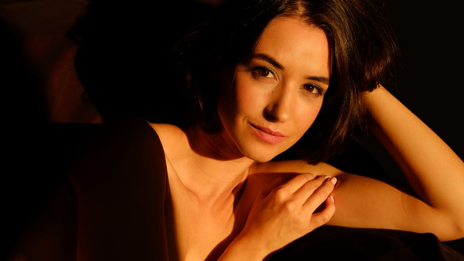 A young woman with dark hair poses in dramatic warm lighting, hands clasped, wearing a black dress.
