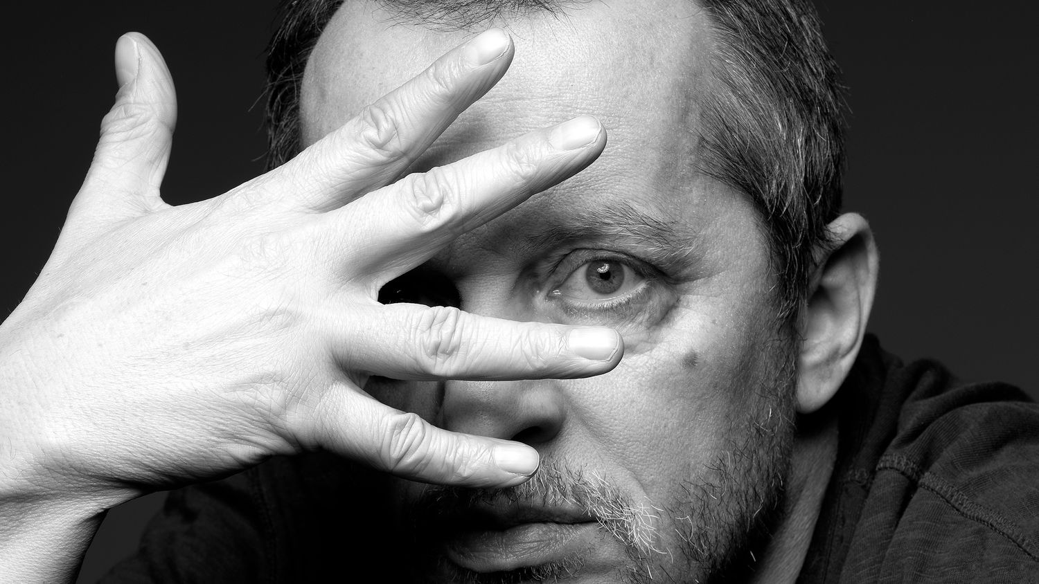 Black and white close-up portrait of a man partially covering his face with his hand, one eye visible.