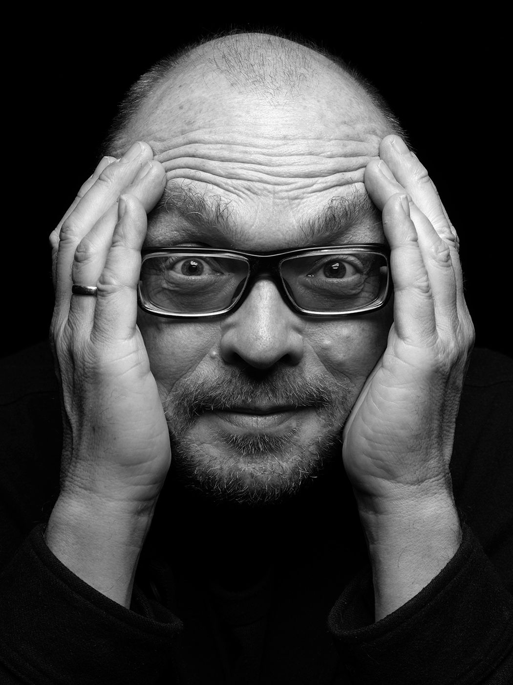 Black and white portrait of a bald man with glasses holding his head in both hands, wide-eyed. Bugge Wesseltoft
