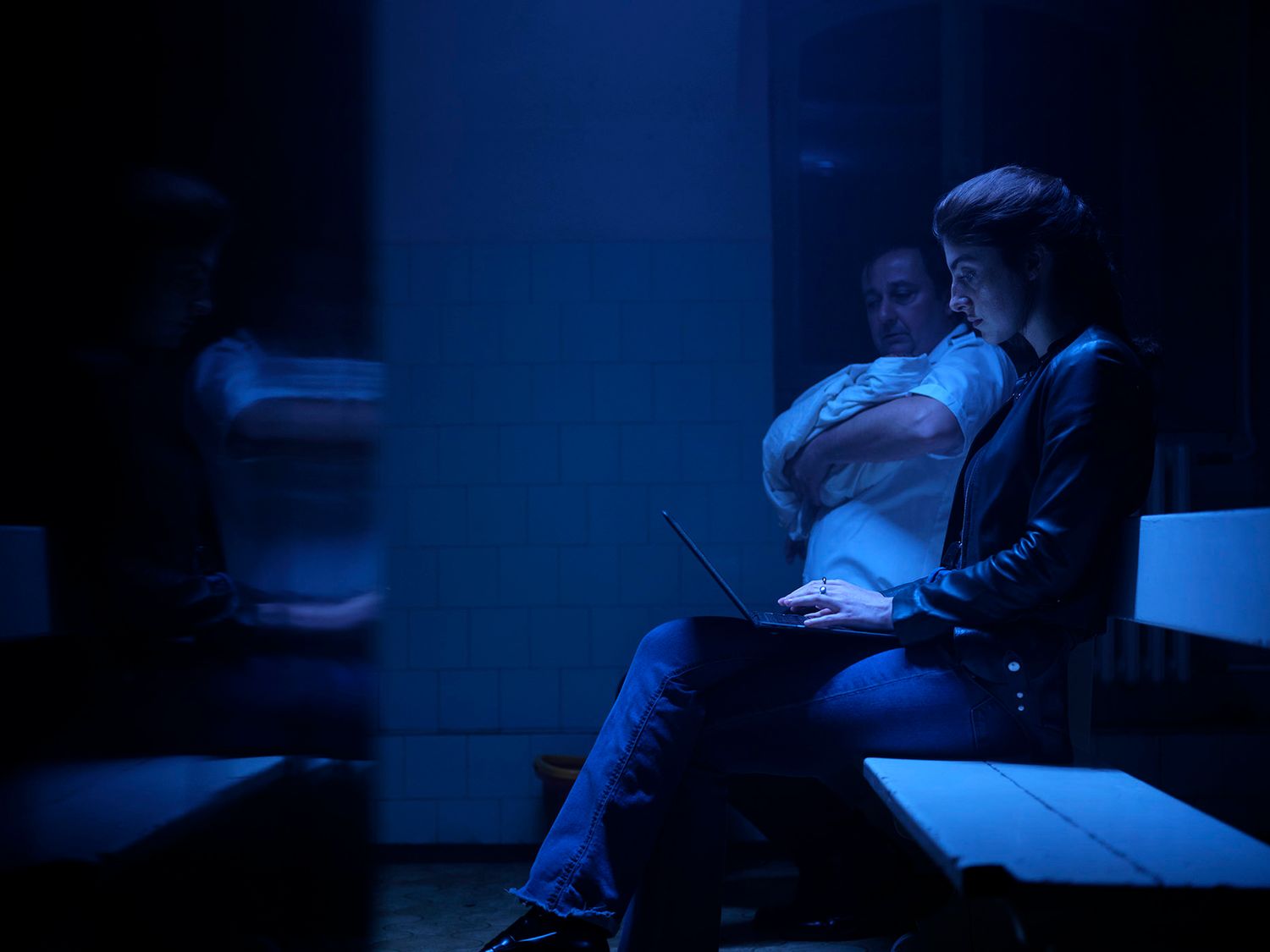 Two people sitting in dark blue-lit room, one using a laptop computer.