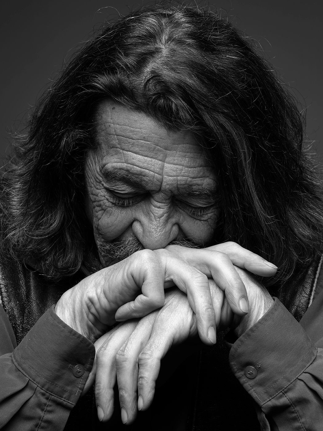 Black and white portrait of an elderly person with long hair, eyes closed, hands clasped near face.