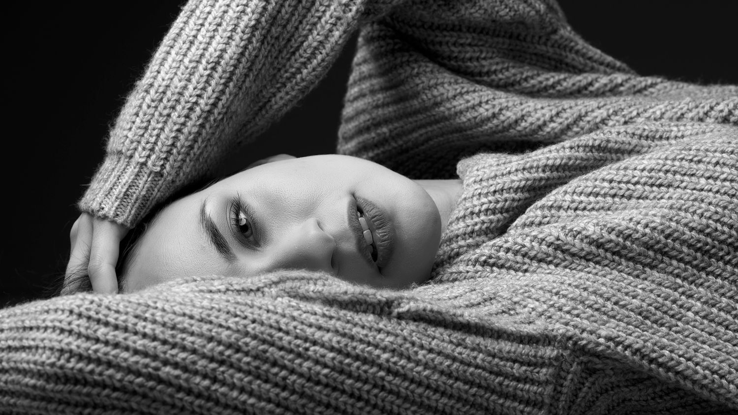 A woman wrapped in a chunky knit sweater lies down in a black and white portrait photo.