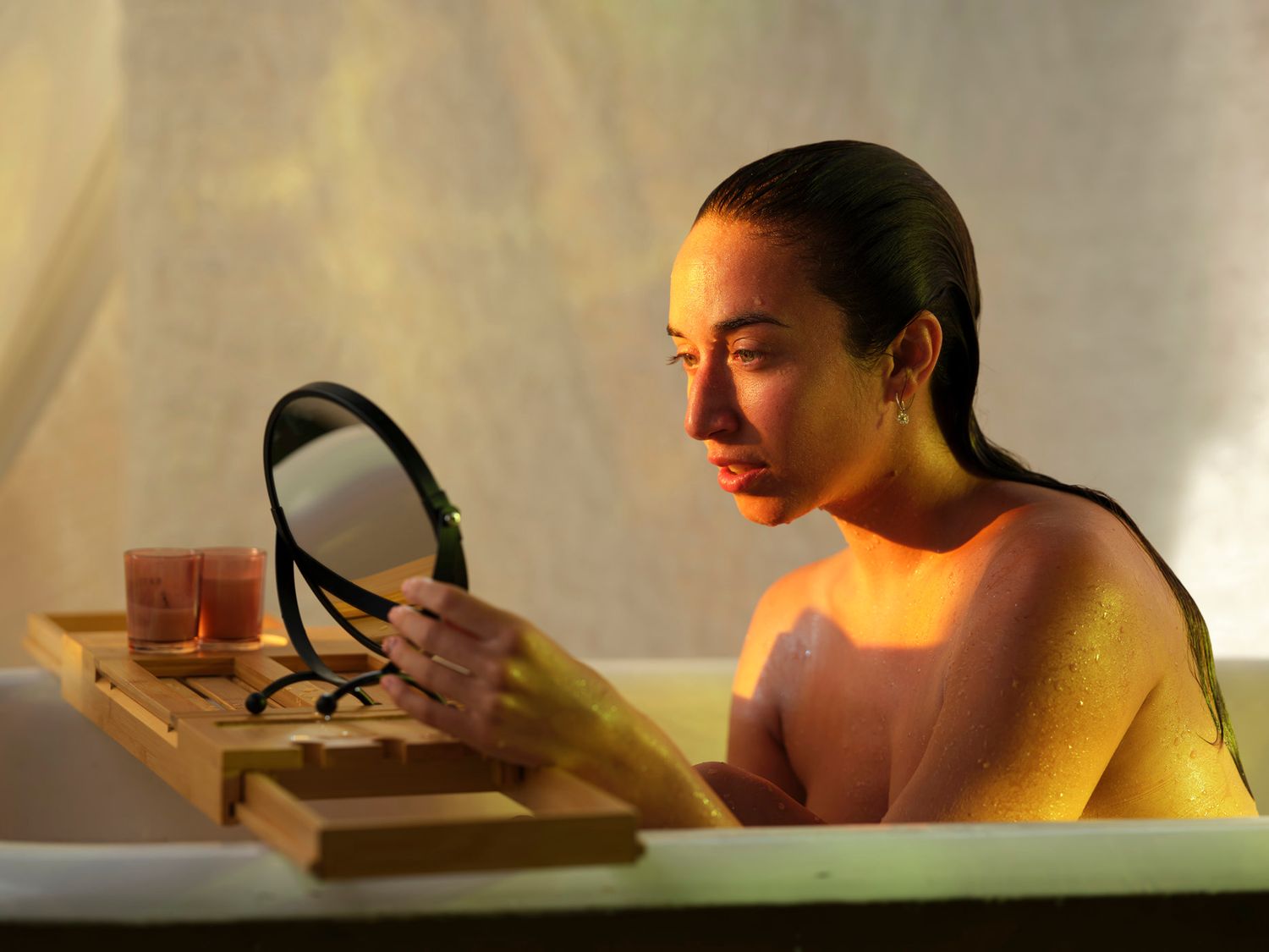 A woman examines her reflection in a handheld mirror while relaxing in a bathtub.