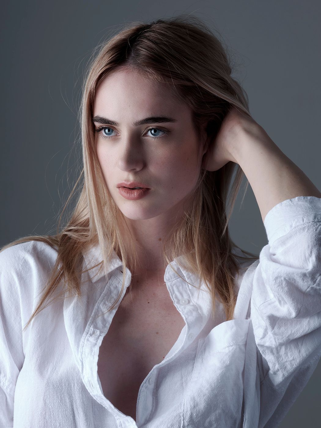 Young woman with blue eyes and blonde hair posing in a white button-up shirt against a gray background.