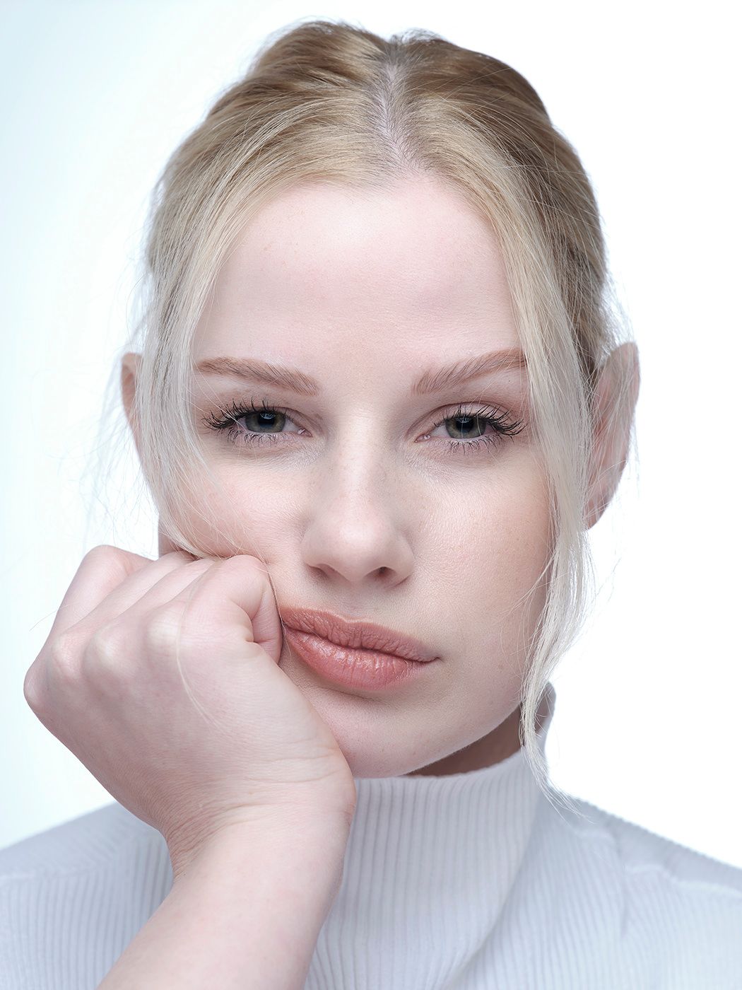 Young blonde woman resting chin on hand, wearing white turtleneck, with a contemplative expression on light background.