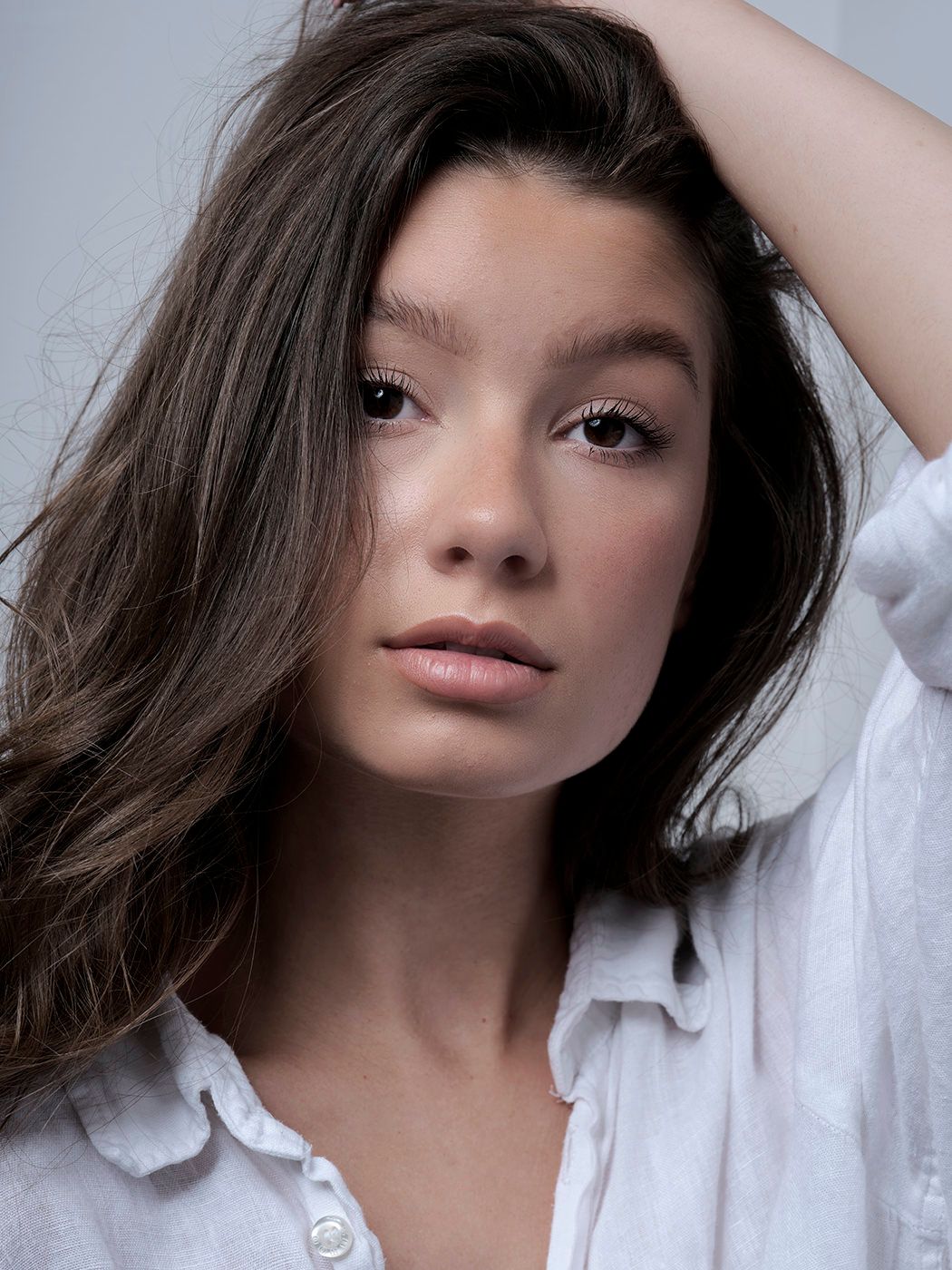 Young woman with dark wavy hair in white linen shirt poses with hand behind head against gray background.