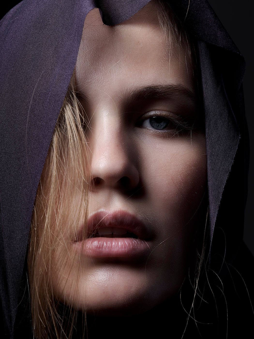 Close-up portrait of a young woman with blue eyes wearing a dark purple hood in dramatic lighting.