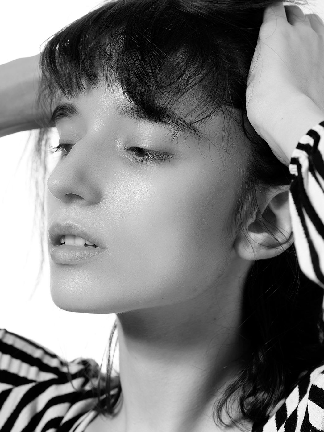 Black and white close-up of a young woman with dark bangs posing in a striped top with hands behind her head.