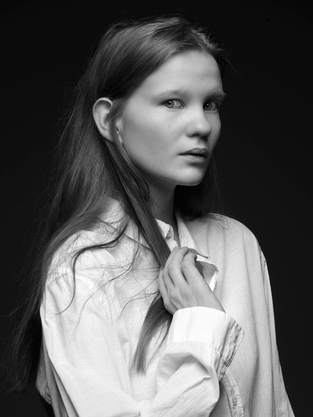 Black and white portrait of a young woman with long hair wearing a white oversized shirt against dark background.