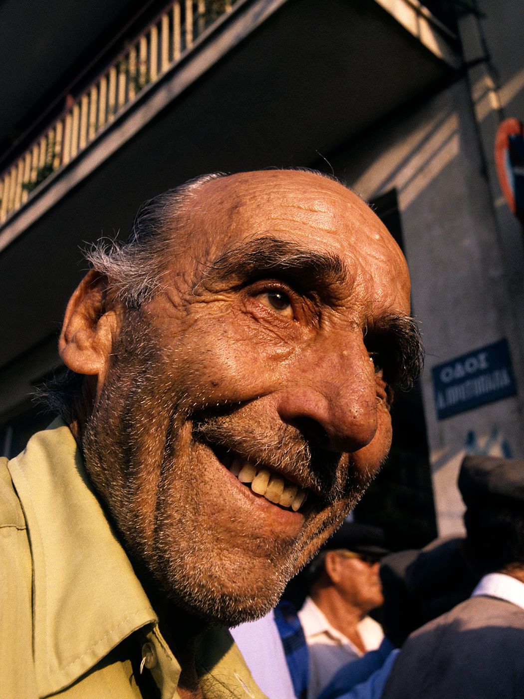 Elderly man with a warm smile, weathered face, and thinning hair photographed outdoors in natural sunlight.