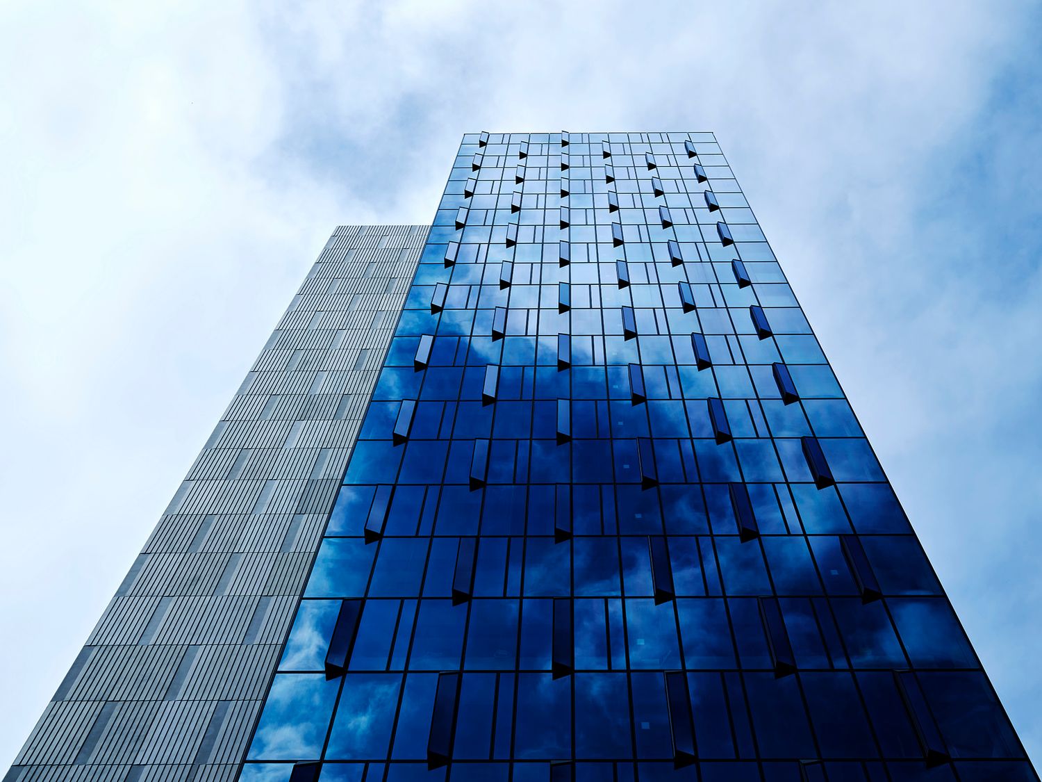 Modern glass skyscraper reflecting blue sky and clouds, viewed from ground level against a cloudy sky.