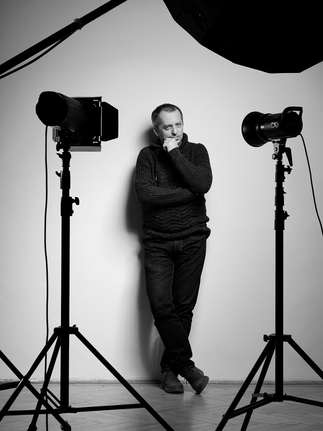 Black and white photo of a man in a dark sweater standing between two studio photography lights on tripods.