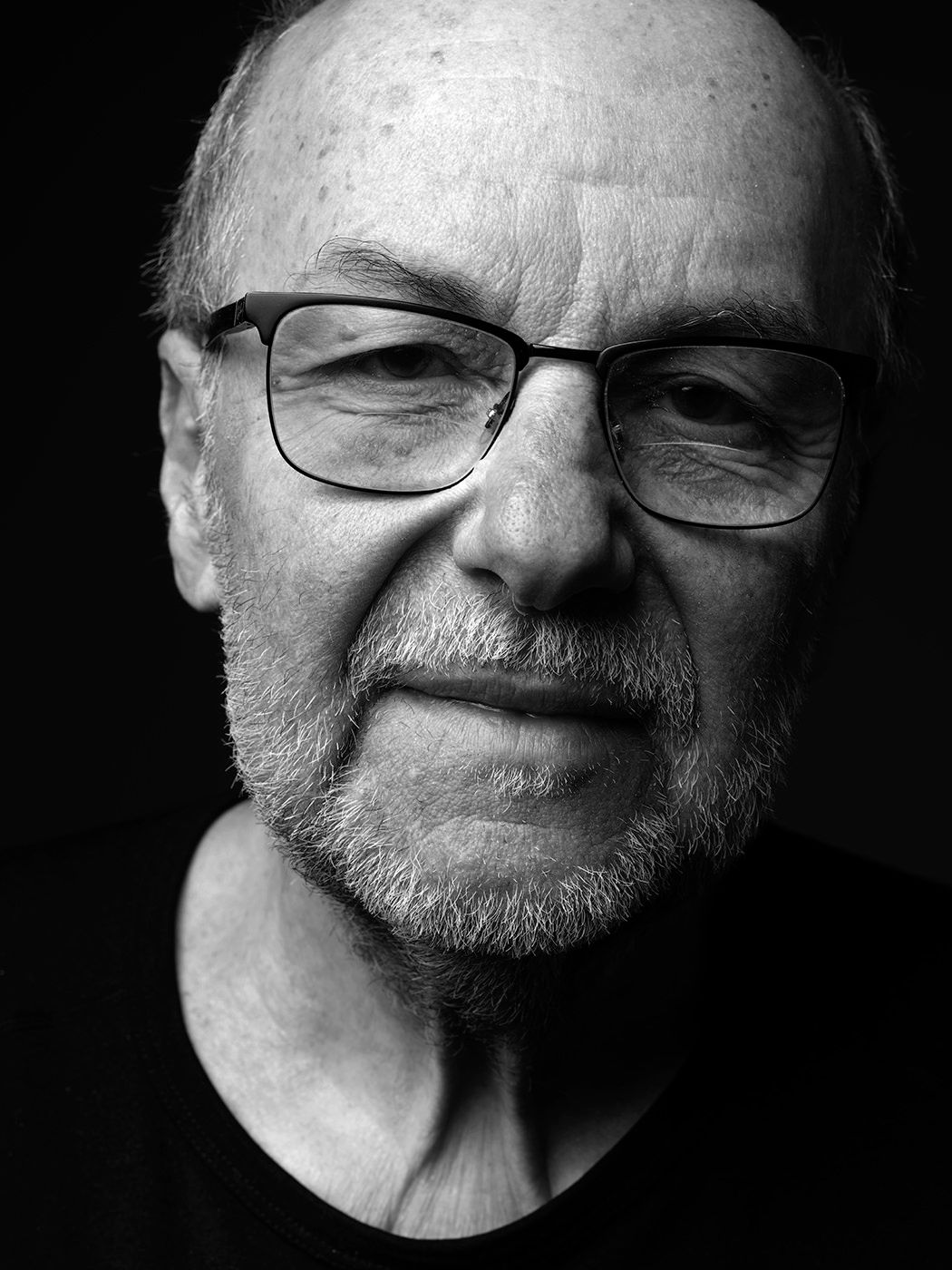 Black and white portrait of an elderly bald man with glasses and a beard against a dark background. László Attila