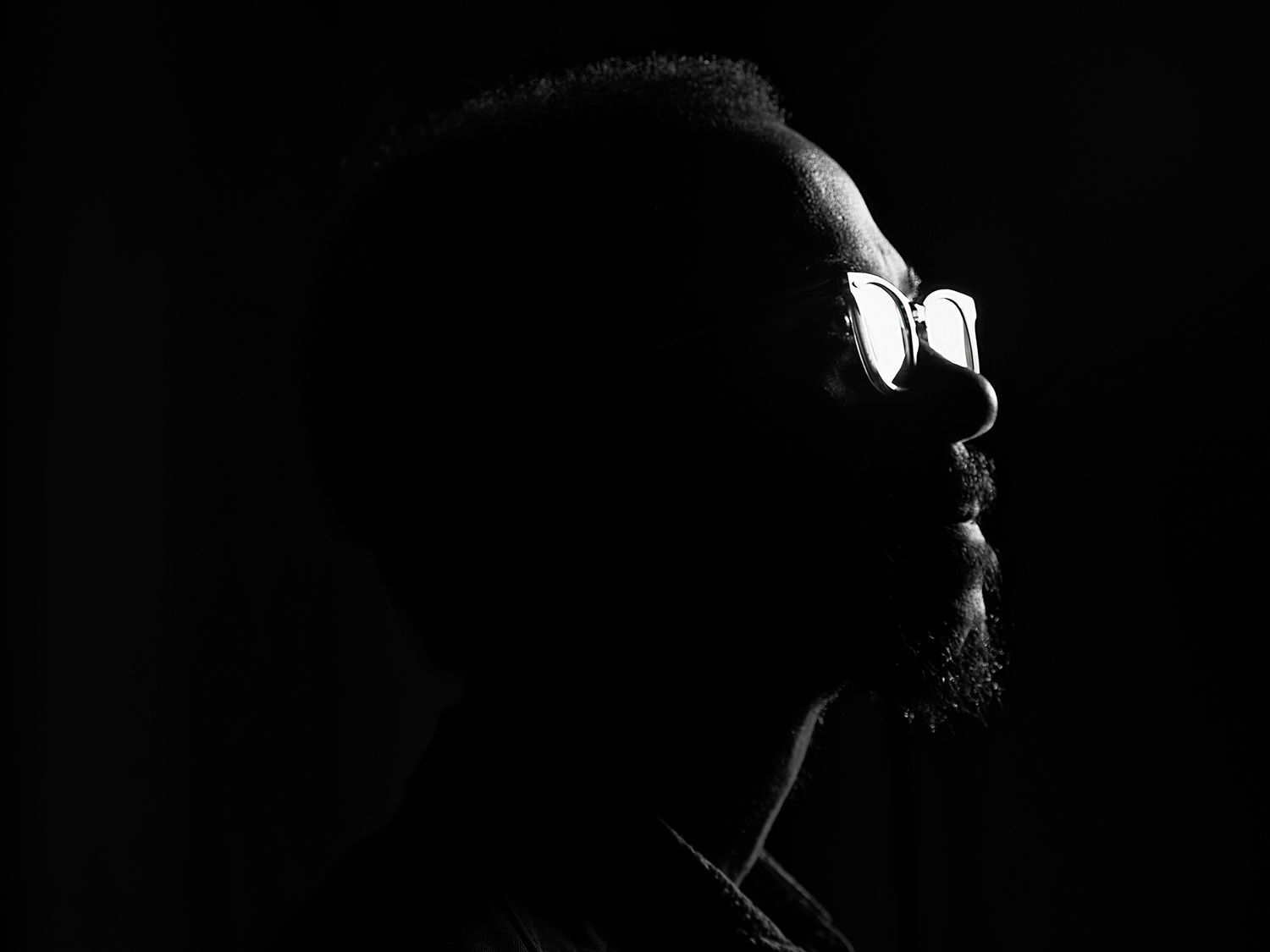 Dramatic black and white profile silhouette of a man wearing glasses against a dark background. Brian Blade