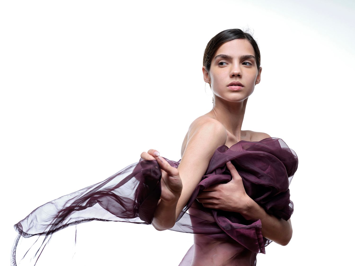 Woman draped in flowing purple sheer fabric against white background, artistic fashion portrait.