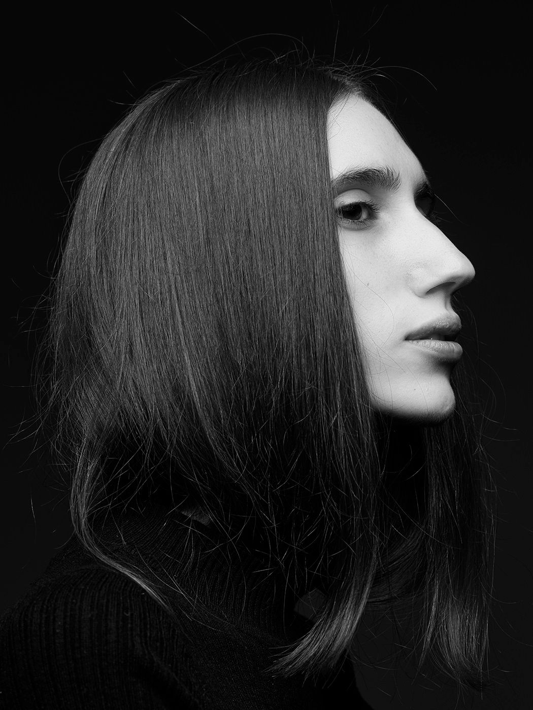 Black and white profile portrait of a young person with long straight dark hair against a dark background.