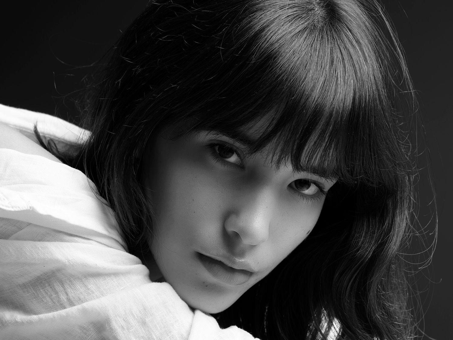 Black and white portrait of a young woman with dark hair and bangs, wearing a white top, looking over her shoulder.