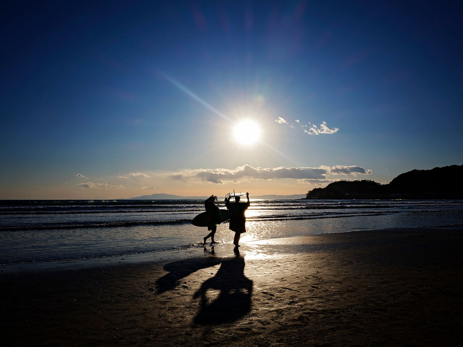 Silhouetted figures on a beach at sunset with sun rays and golden reflections on wet sand.