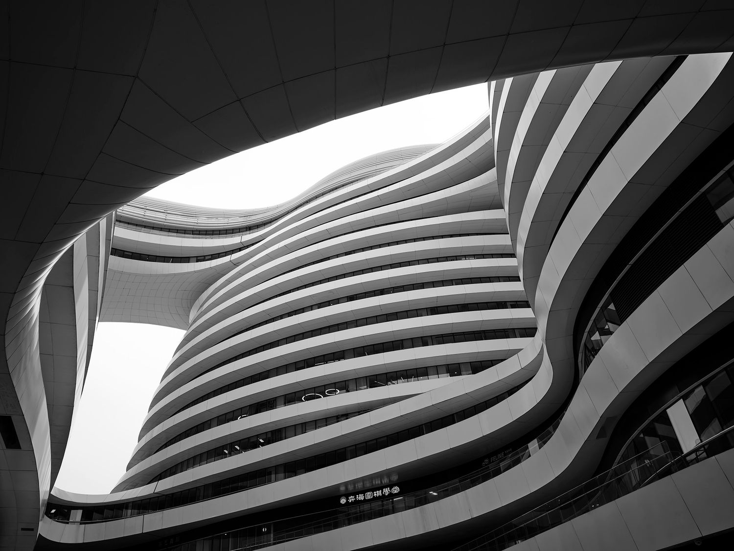 Black and white photo of a futuristic curved building interior with layered wavy floors and an open skylight above.
