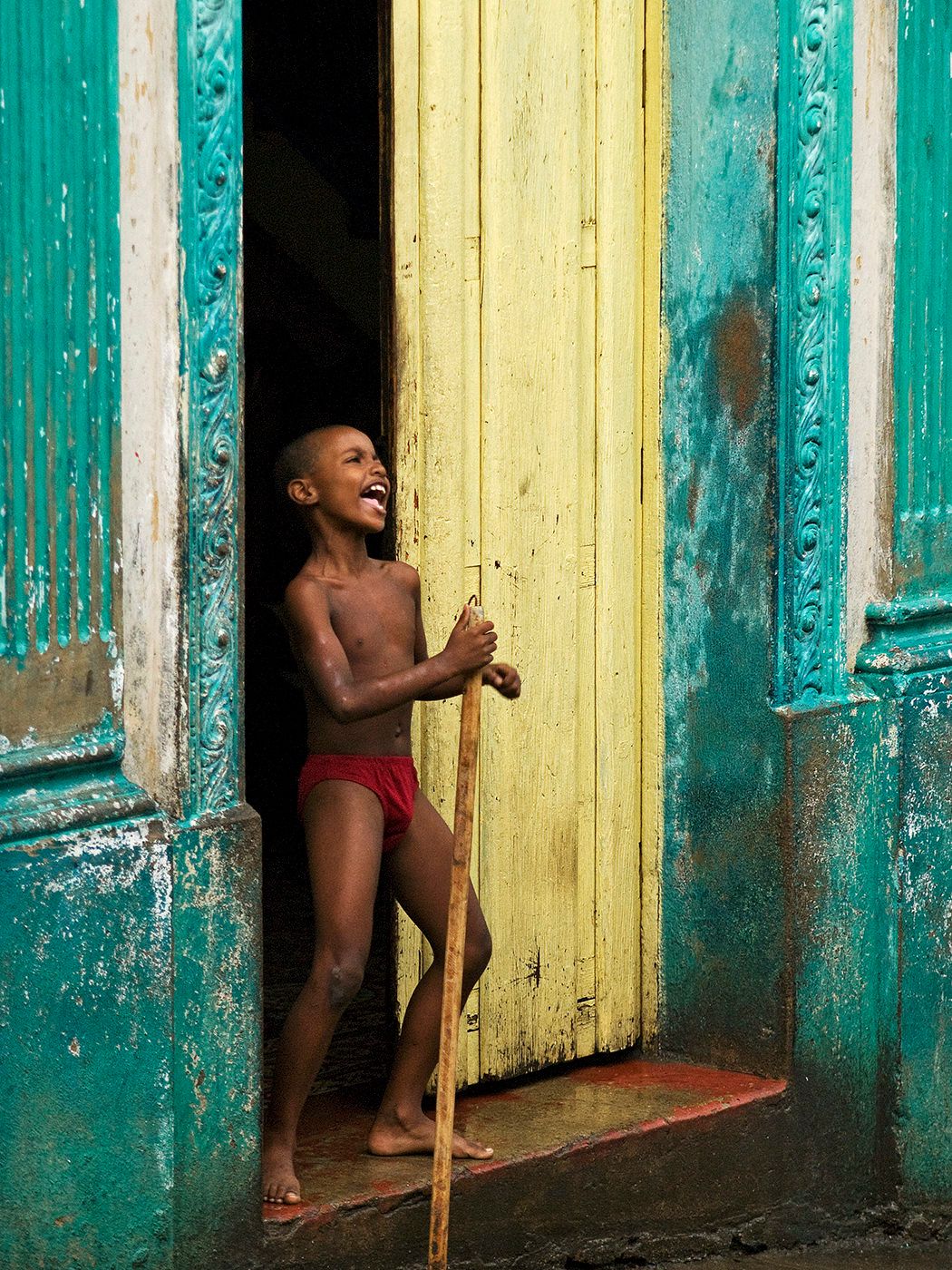 Joyful young boy laughing in a doorway, holding a stick, framed by vibrant turquoise and yellow painted walls.