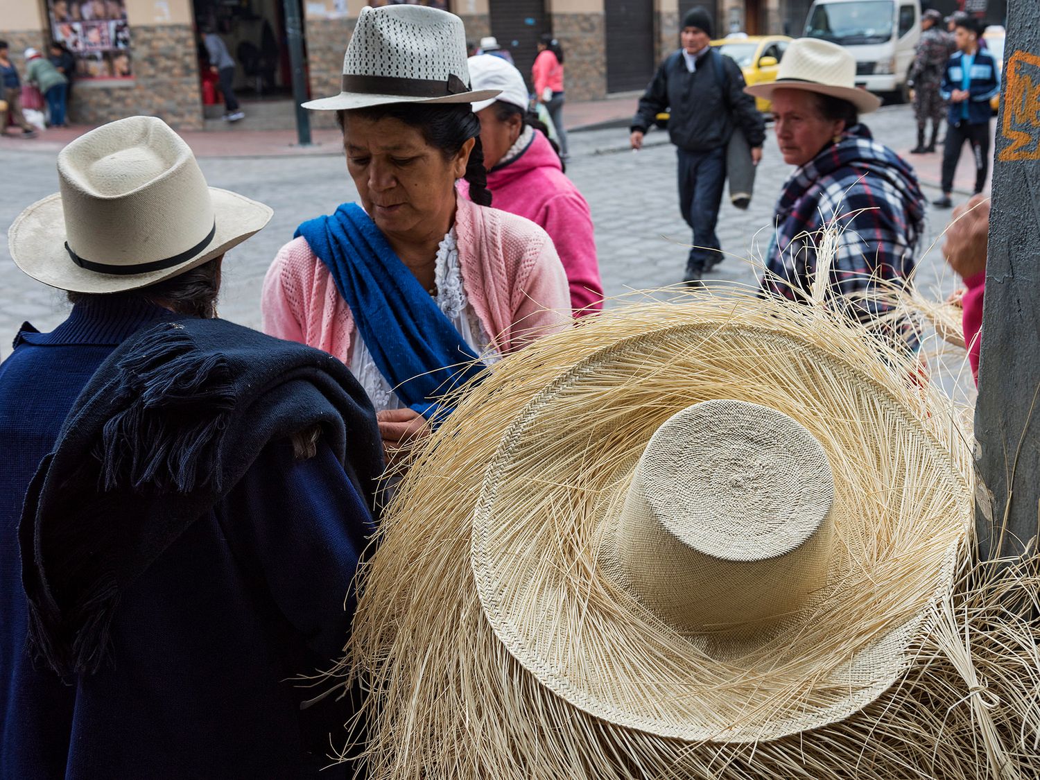 Street market vendors selling traditional straw hats in Ecuador, with locals wearing fedoras in an outdoor setting.