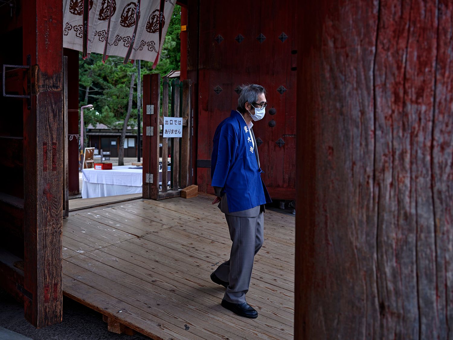 Elderly man wearing blue jacket and face mask walking through traditional red wooden Japanese temple gate.
