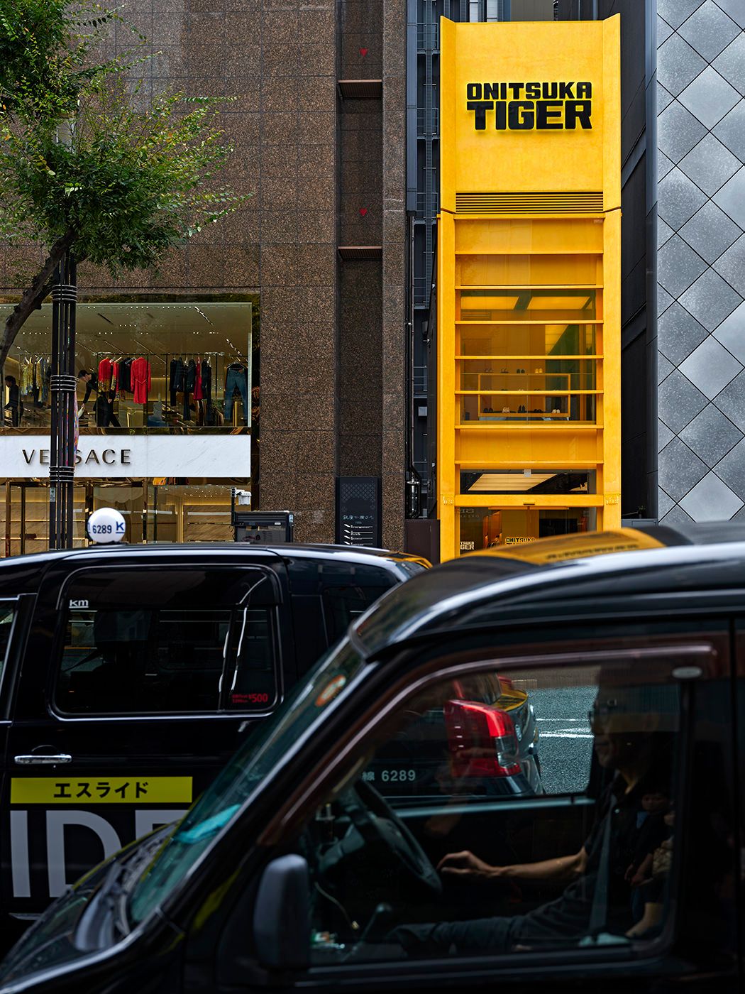 Onitsuka Tiger store with bold yellow facade beside Versace boutique on a busy Tokyo street with black taxis.