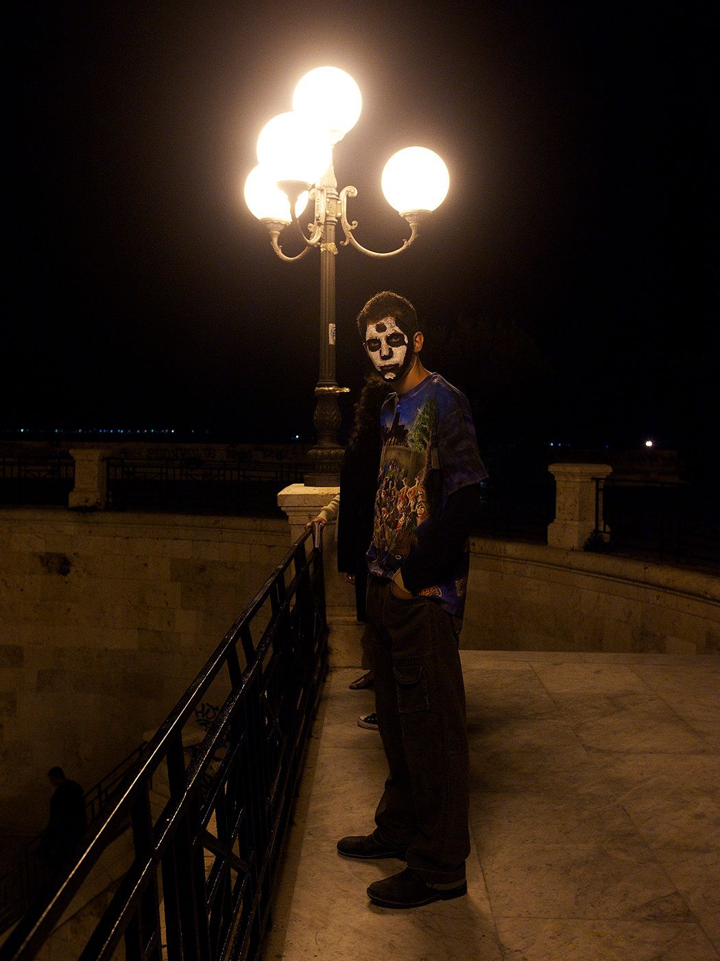 Person in Halloween skeleton face paint stands under glowing street lamp at night on an outdoor terrace.