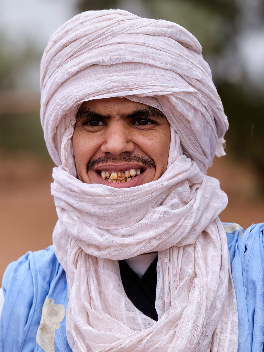 Smiling Tuareg man wearing a traditional white turban and blue robe in a desert setting.