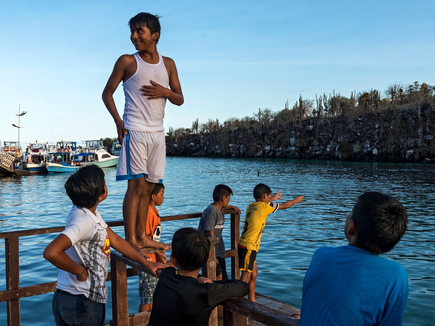 Children playing on a wooden dock over calm blue water with boats and rocky cliffs in the background.