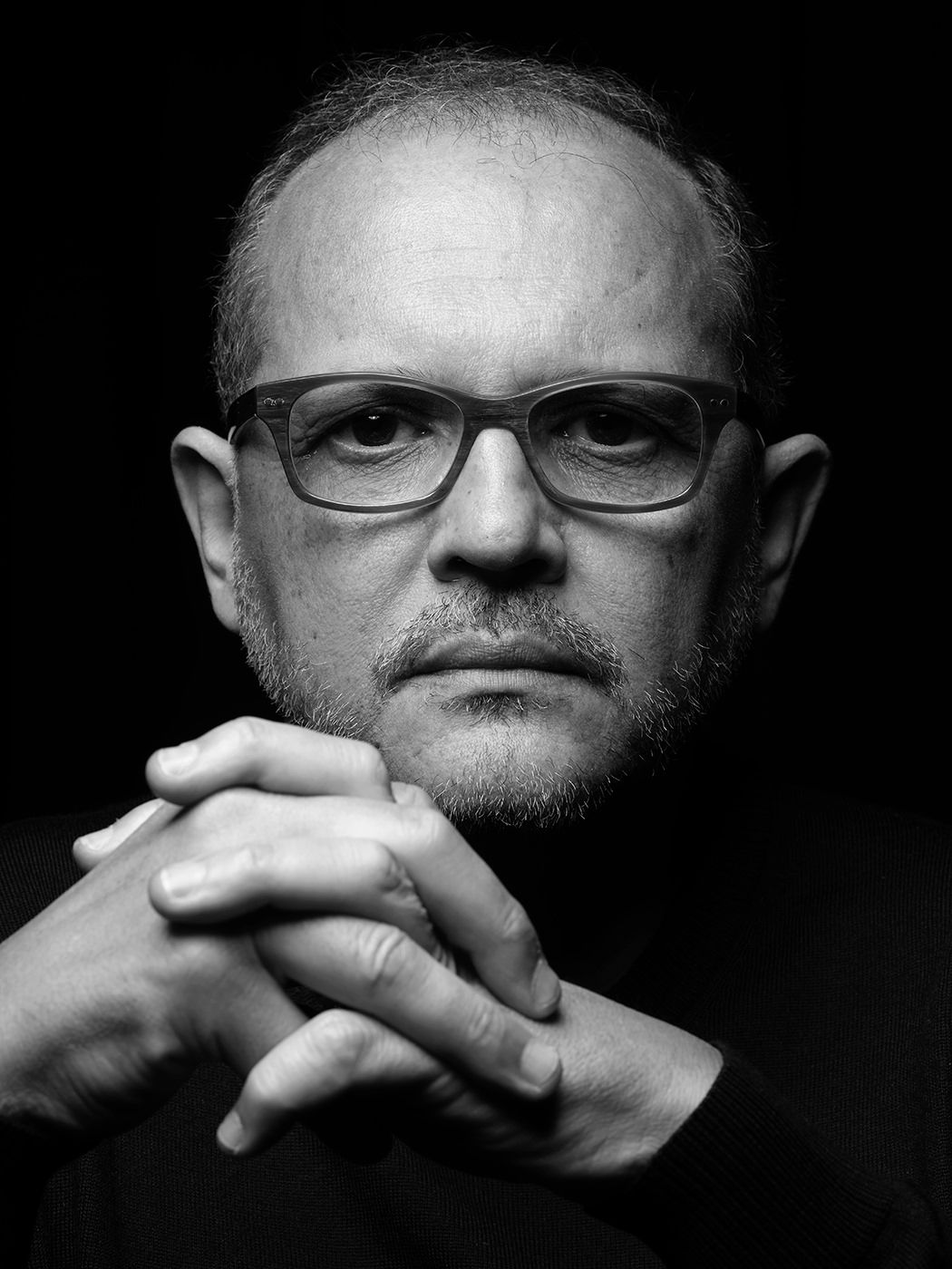 Black and white portrait of a middle-aged man with glasses, clasped hands, and a serious expression against dark background. Anouar Brahem