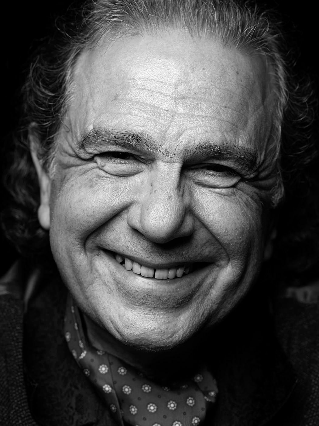 Black and white portrait of a smiling older man with wavy gray hair wearing a patterned shirt. Rabih Abou-Khalil, musician.