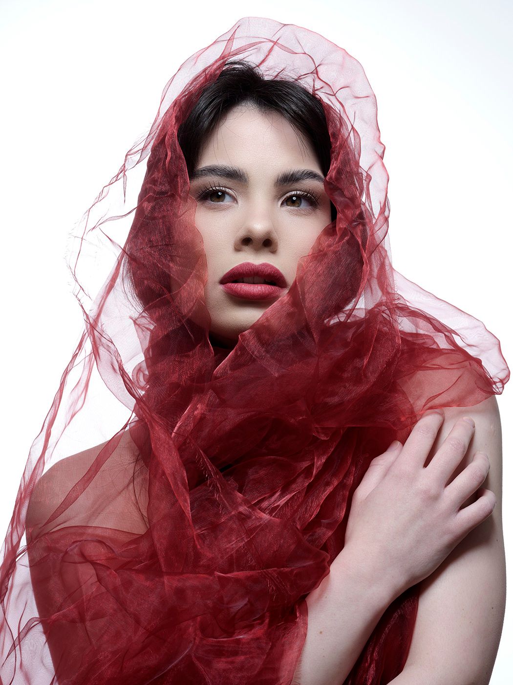 Woman draped in flowing red sheer fabric veil, wearing bold red lipstick against white background.