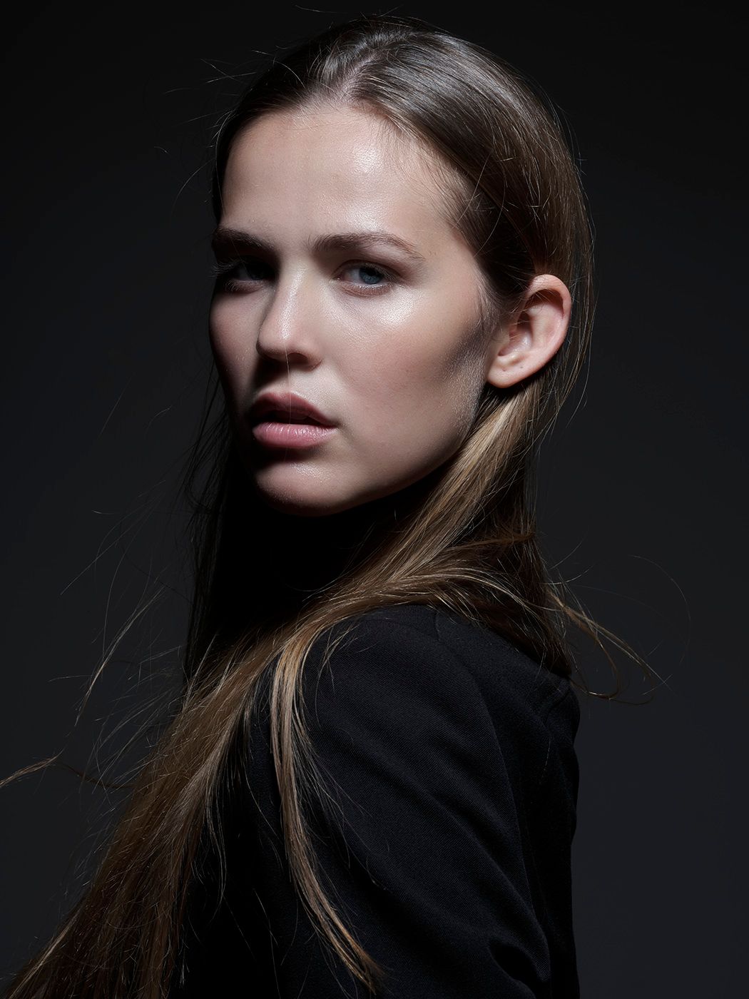 Young woman with long brown hair in black jacket poses against dark background in dramatic studio portrait.
