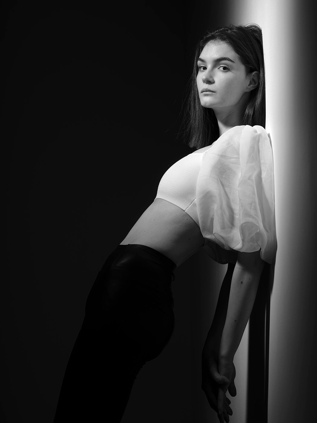 Black and white fashion portrait of a young woman in sheer puff-sleeve crop top leaning against a lit wall.