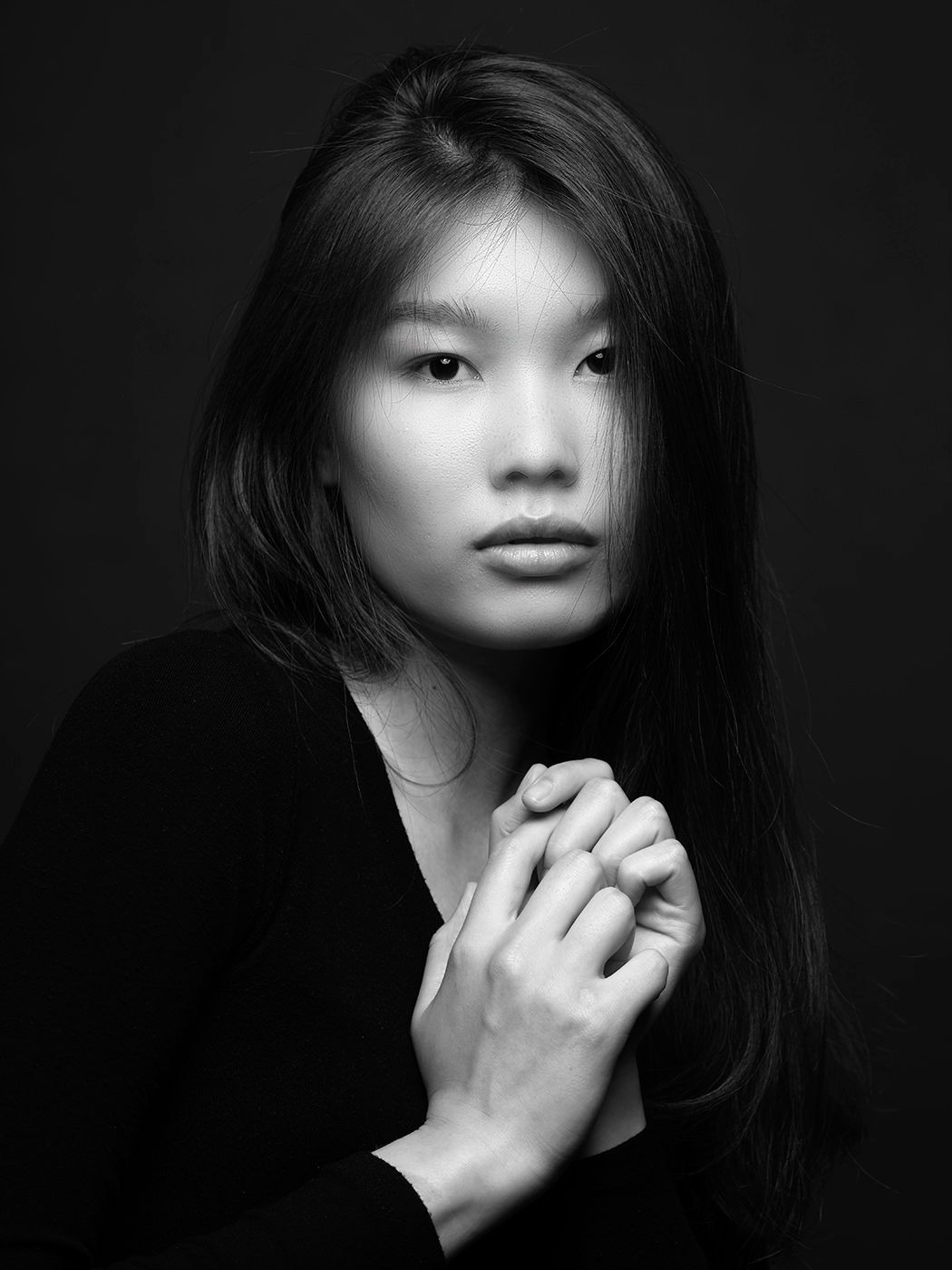 Black and white portrait of a young Asian woman with long dark hair, hands clasped, wearing a black top.