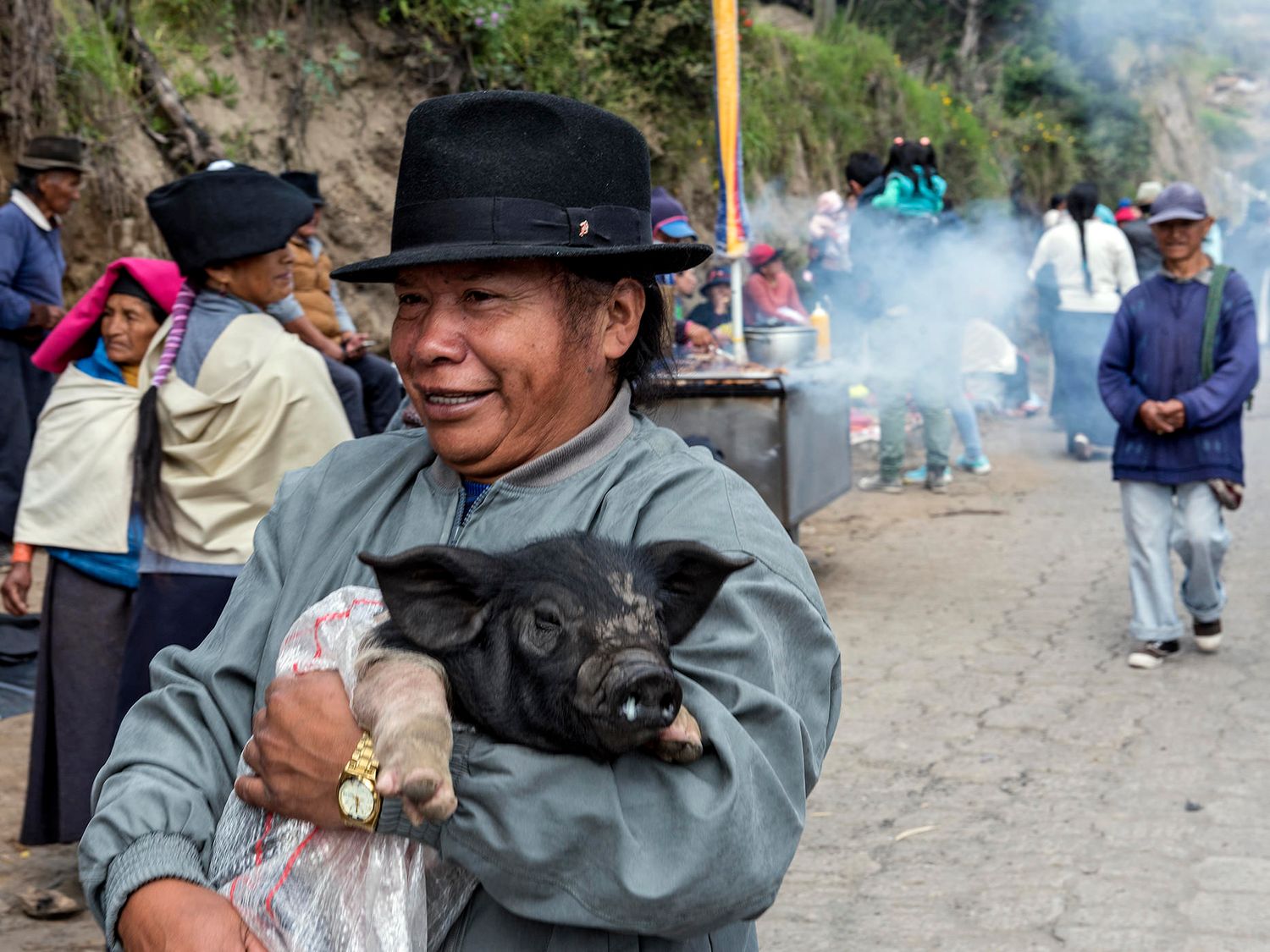 Man in black hat smiling while holding a small black piglet at a busy outdoor market in Ecuador.