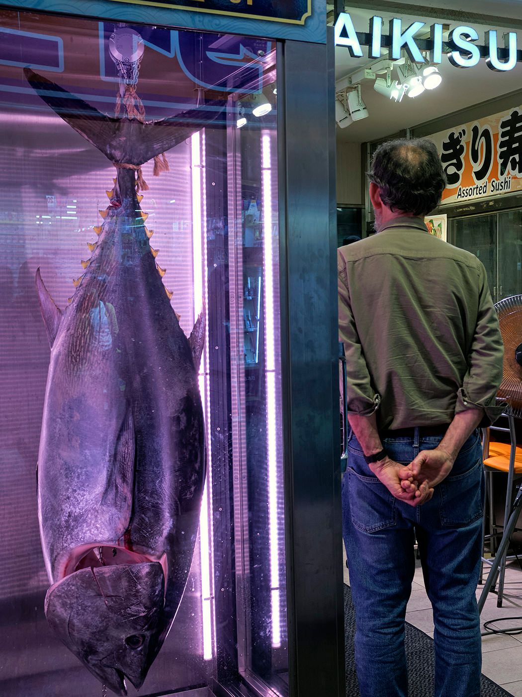 A man stands before a large whole tuna fish displayed in an illuminated case outside a Japanese sushi restaurant.