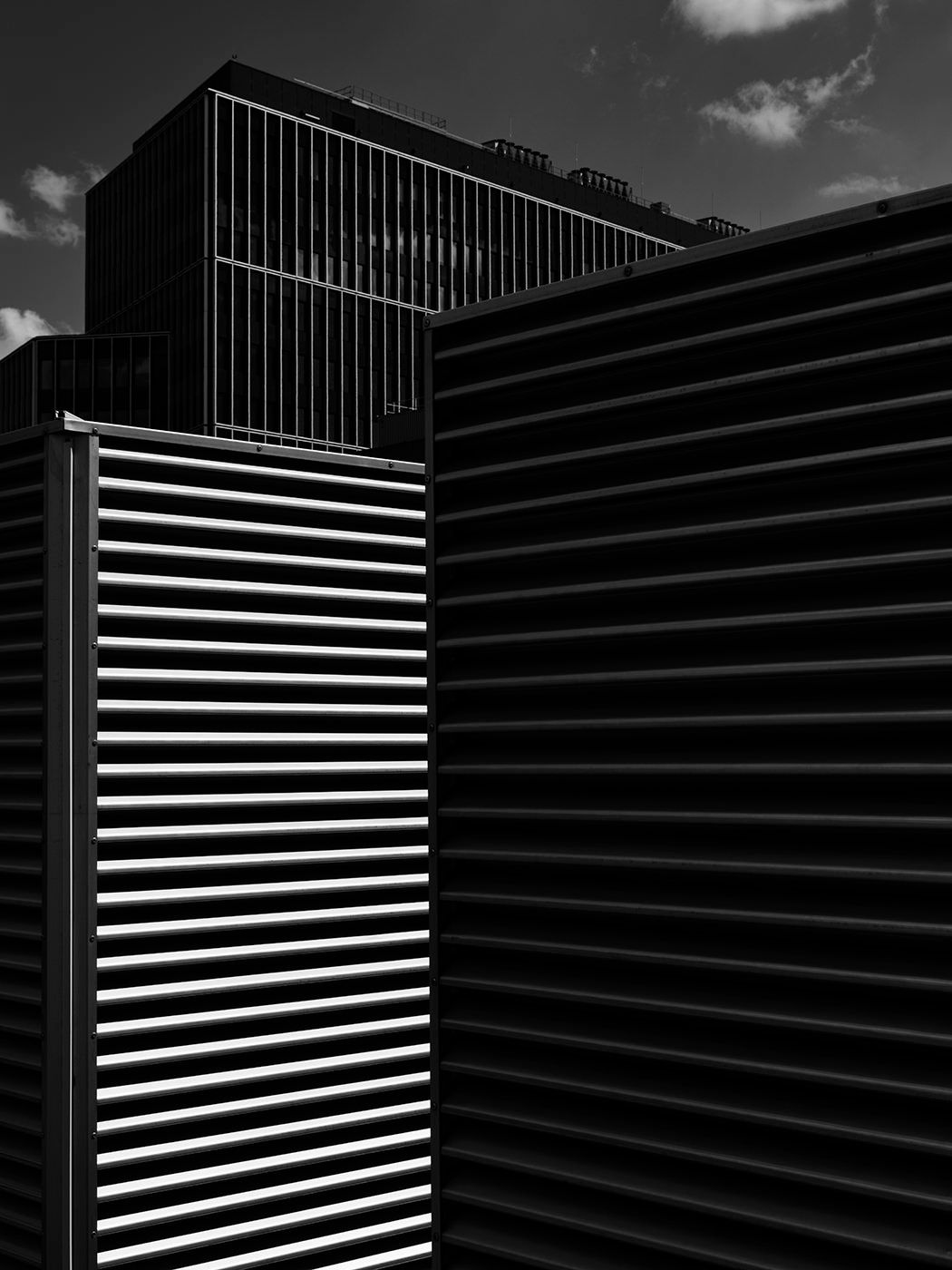 Black and white architectural photo of modern buildings with horizontal louvers against a cloudy sky.