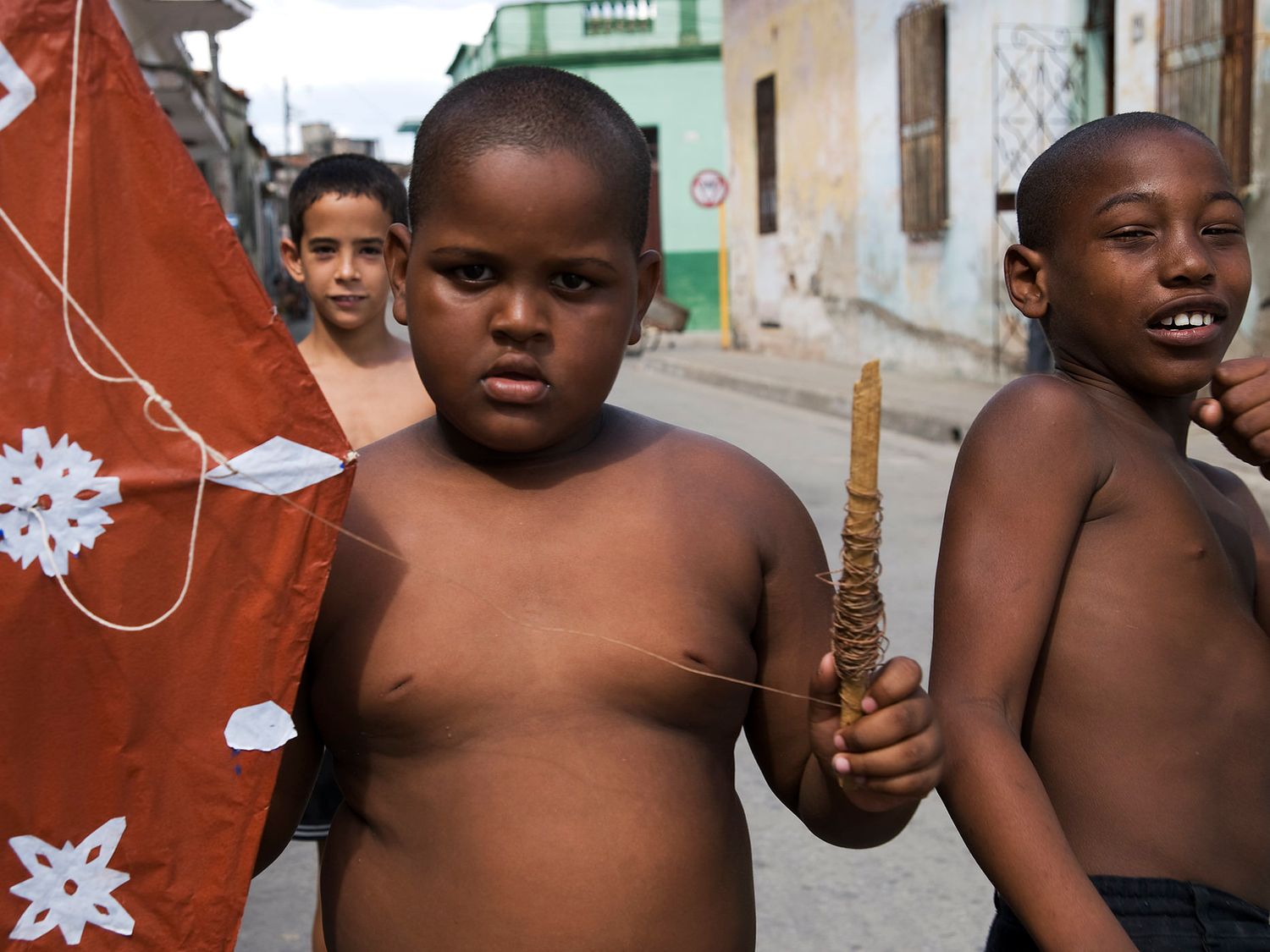 Three shirtless children playing on a street, with one holding a stick and another carrying a red kite.