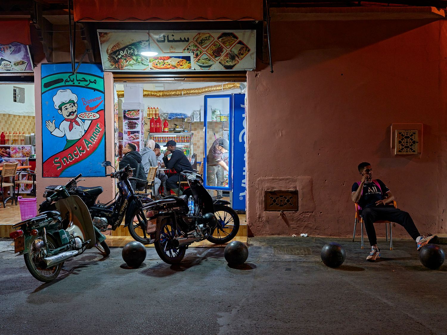 Motorcycles parked outside a vibrant Middle Eastern street food restaurant at night with a man relaxing nearby.