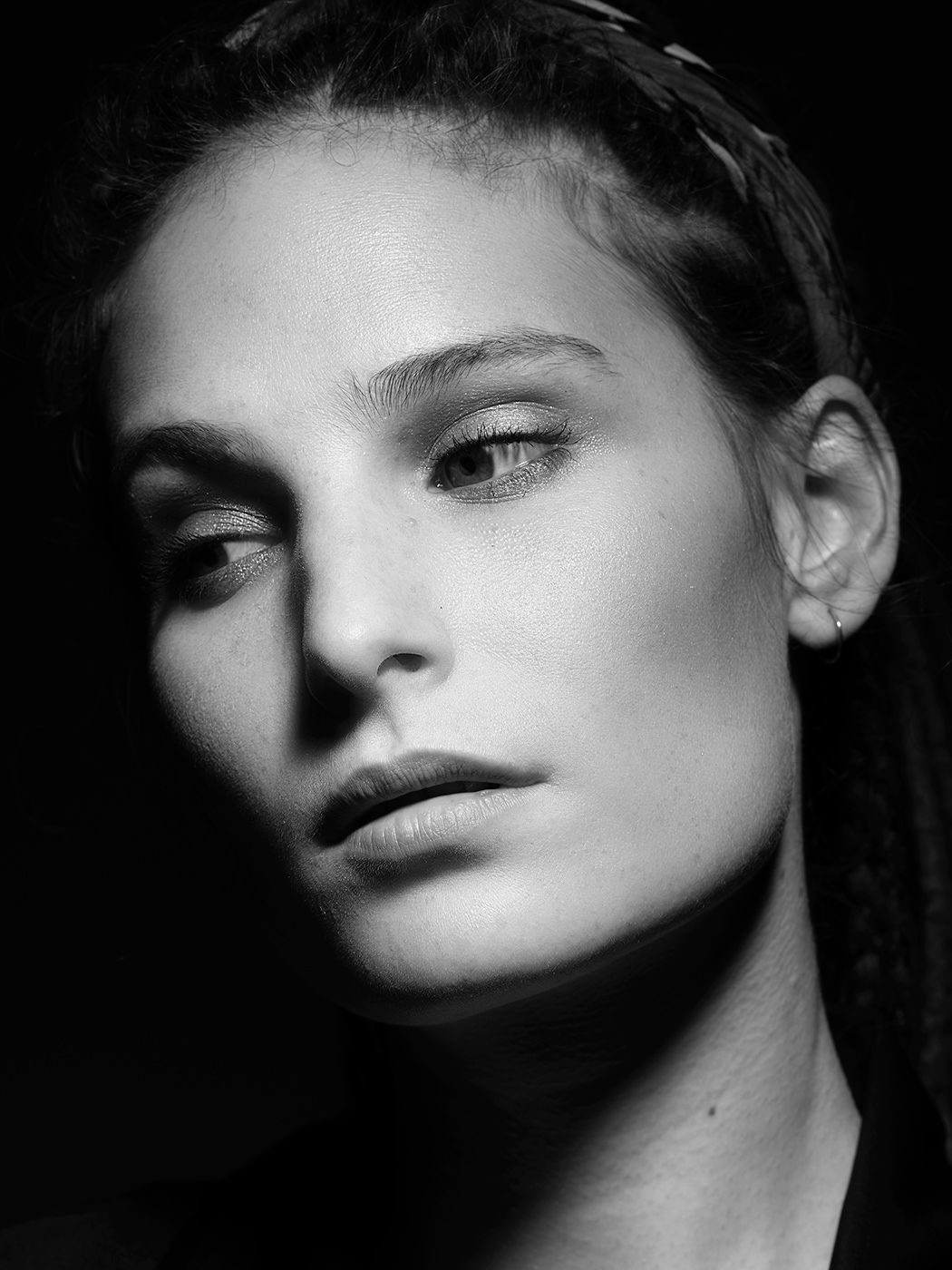 Black and white portrait of a young woman with braided hair and smoky eye makeup against a dark background.