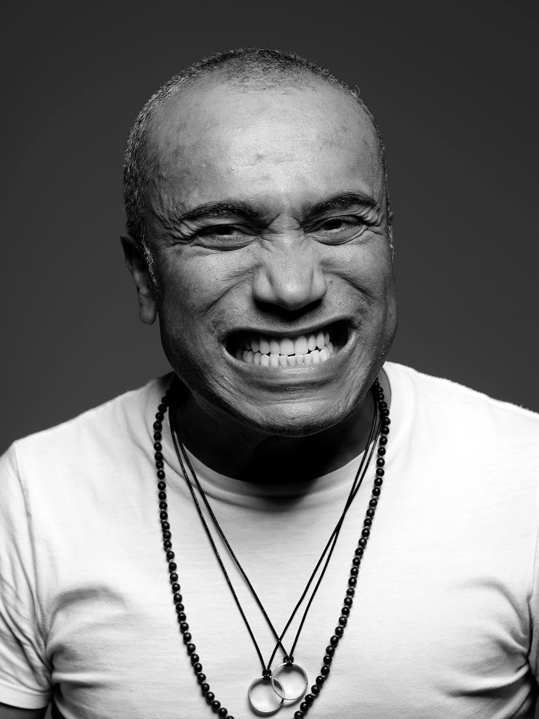 Black and white portrait of a bald man laughing broadly, wearing a white t-shirt and layered bead necklaces. Ernesttico