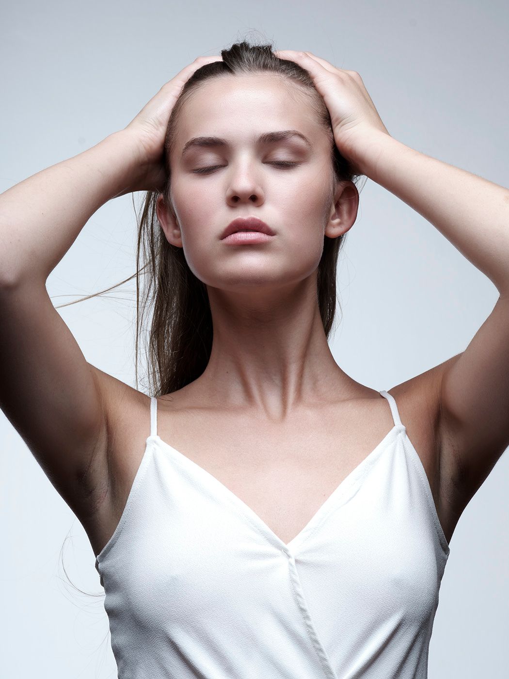 Young woman in white camisole with eyes closed, hands on head, against light gray background.