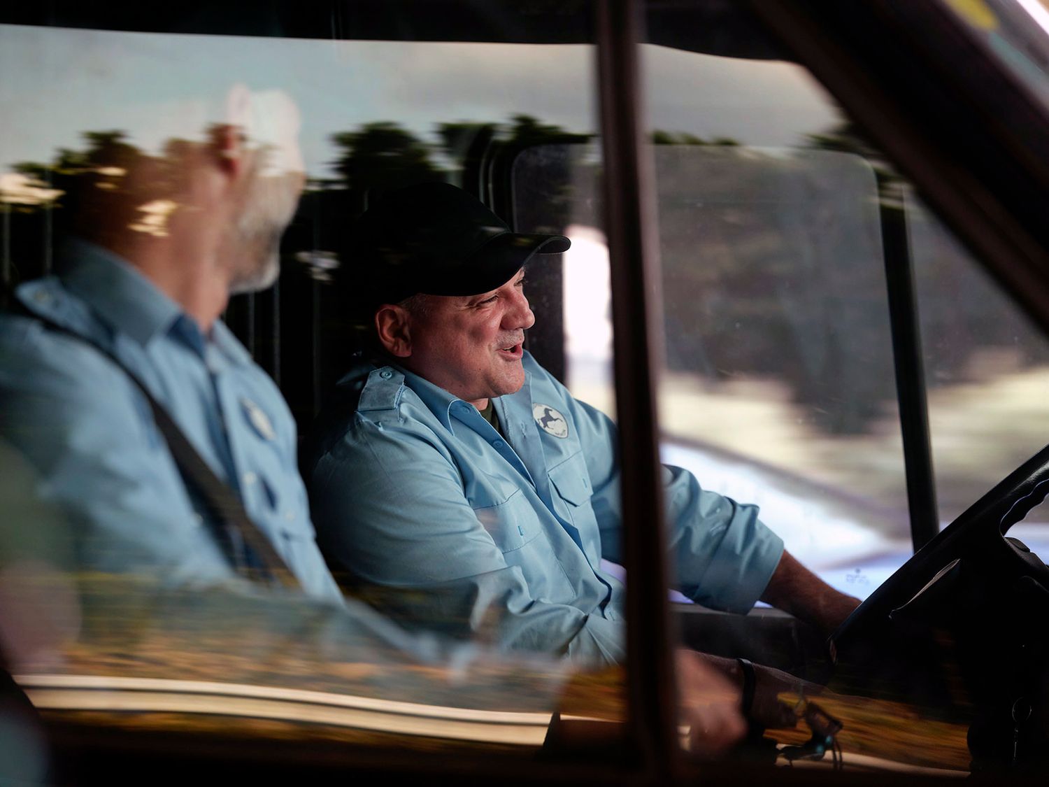 Two men in blue uniforms sit inside a vehicle cab, smiling and conversing through a window reflection.