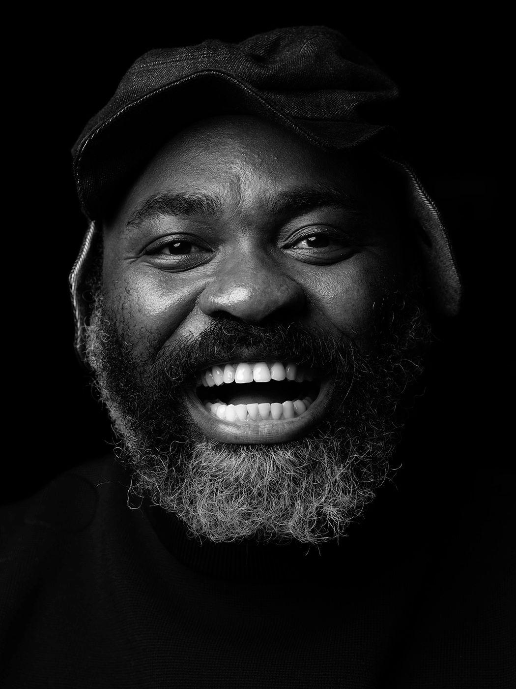 Black and white portrait of a laughing bearded man wearing a flat cap against a dark background. Nduduzo Makhathini