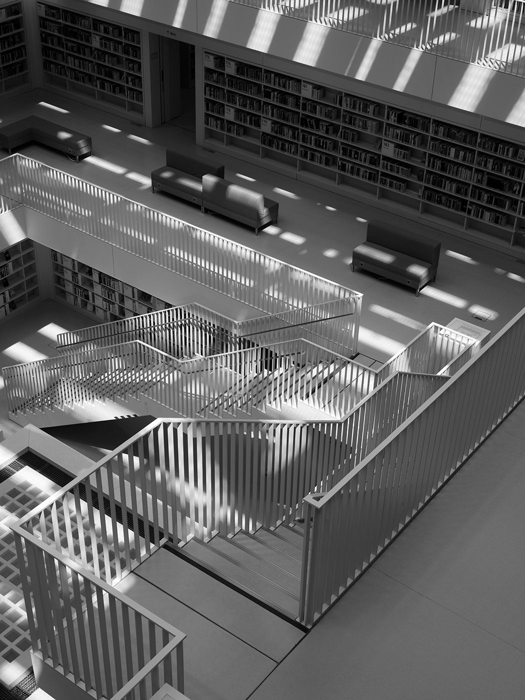 Black and white aerial view of a modern library staircase with geometric railings, bookshelves, and dramatic light patterns.