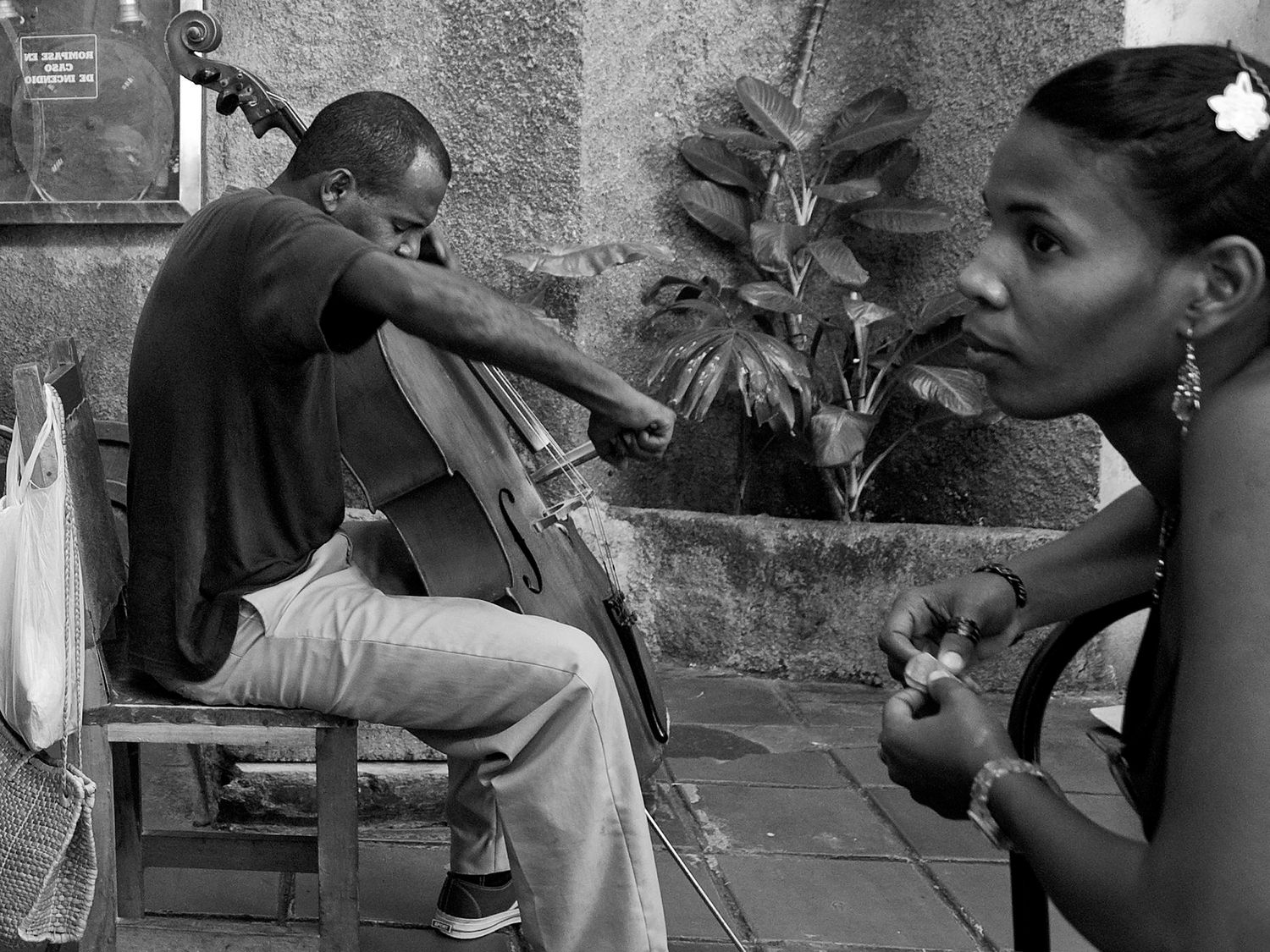 Black and white photo of a man playing cello on a bench while a woman watches nearby on a city street.