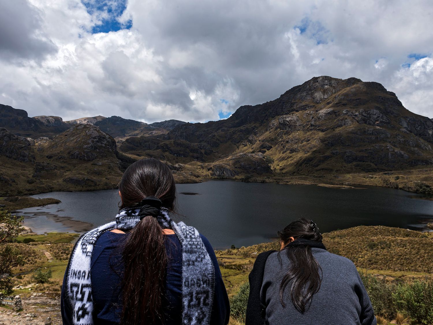 Two women overlook a scenic mountain lake under cloudy skies in a rugged highland landscape.