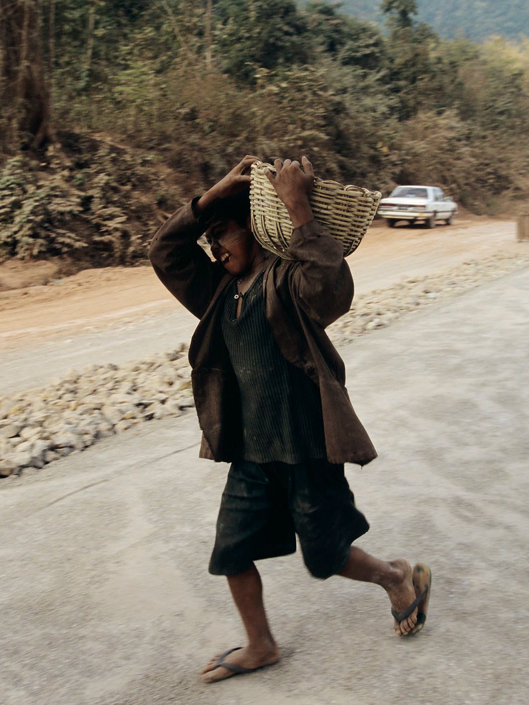 Child carrying woven basket on head while walking along rural road in traditional clothing.